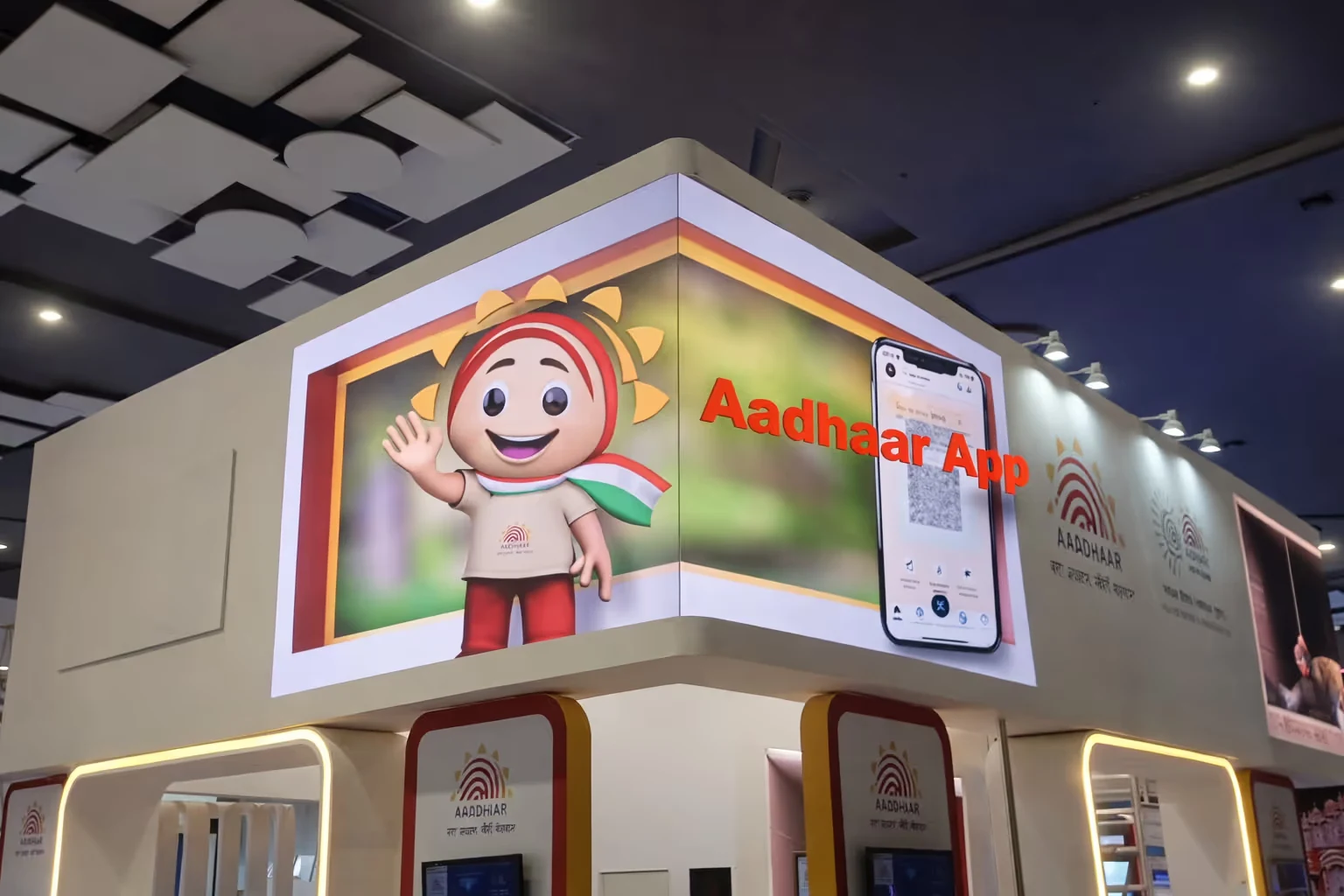 A corner display at an event featuring a 3D cartoon mascot with a sun hat and an Indian tricolour scarf waving, alongside an image of a mobile phone showing the Aadhaar app interface.