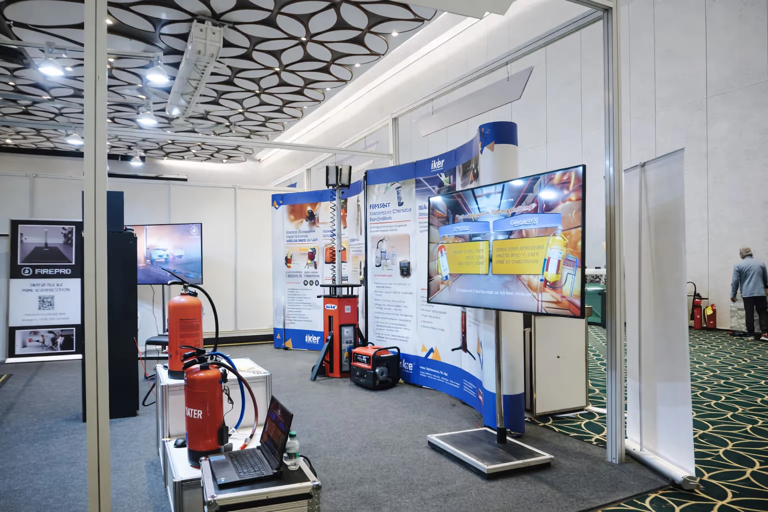 Exhibition booth displaying fire safety equipment including fire extinguishers, a laptop, informational banners, and a large screen TV in a conference hall.