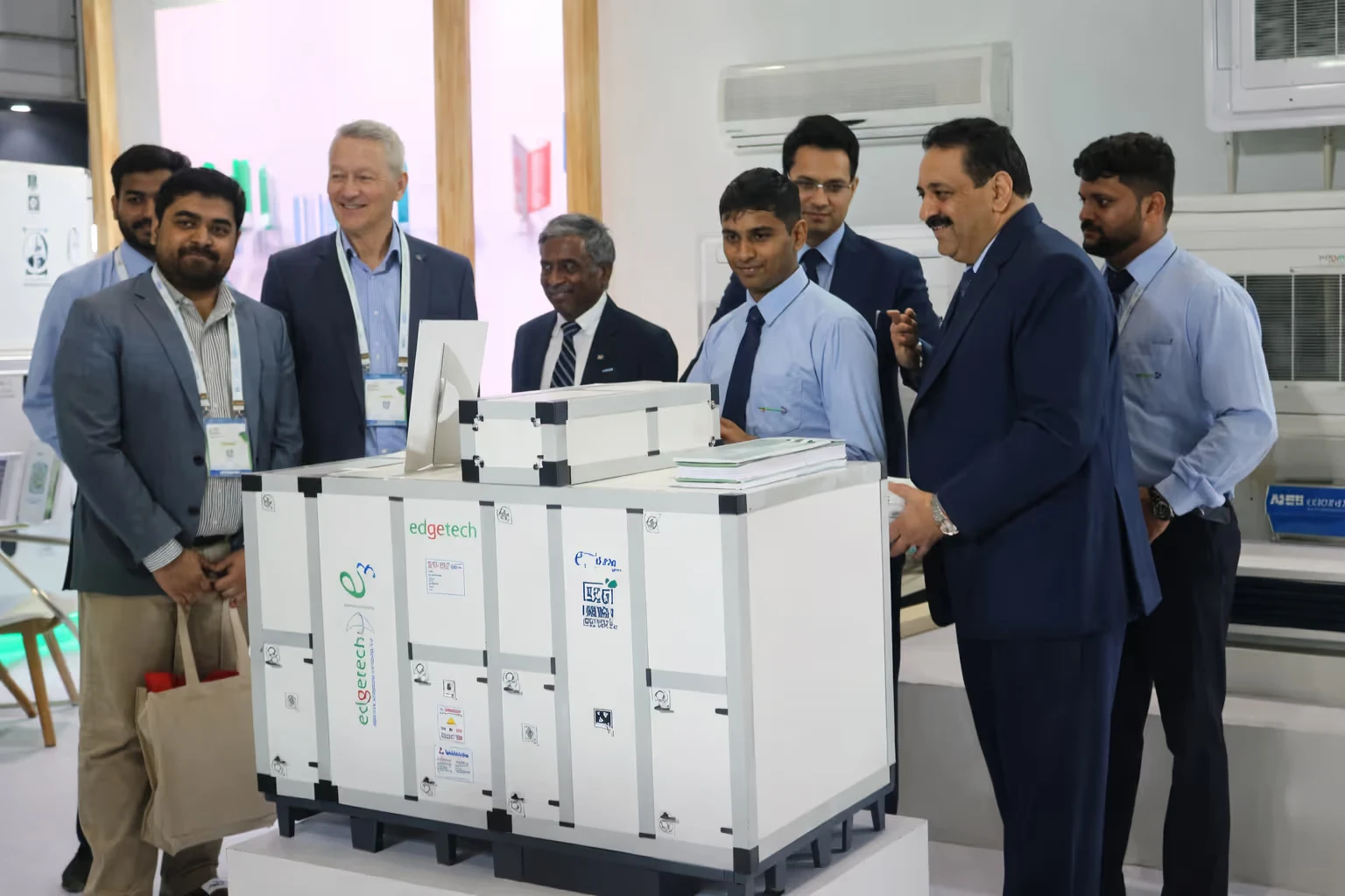 Group of men in business attire standing around a large white industrial device labeled edgetech at an exhibition or trade show.