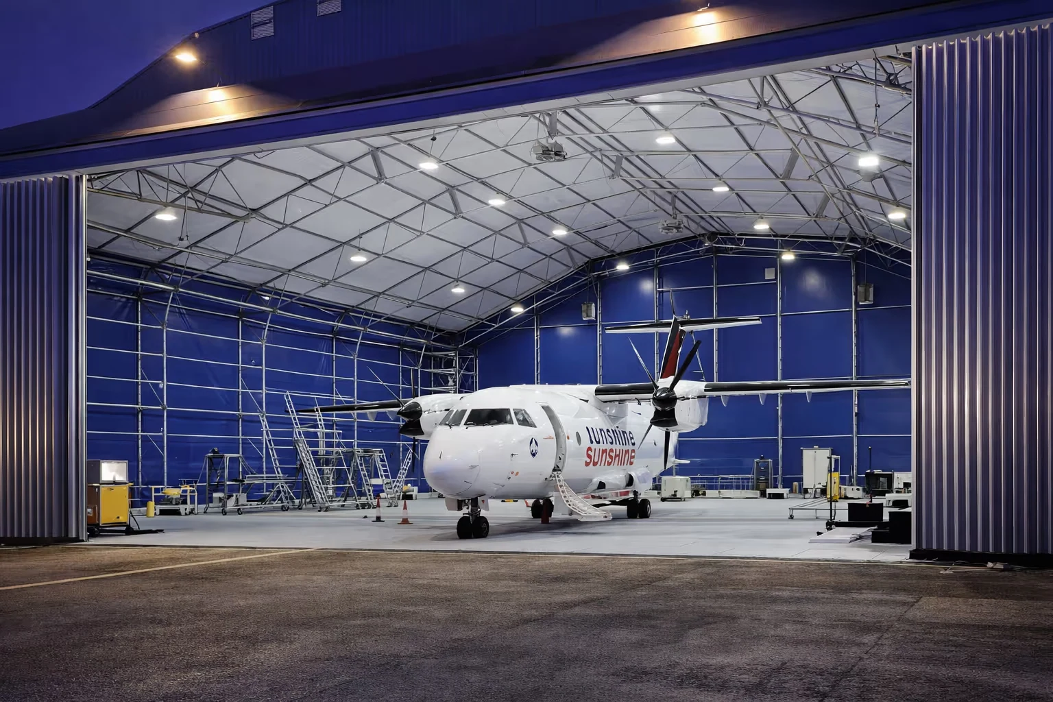 White propeller airplane with 'Sunshine' branding parked inside a large blue-lit hangar at dusk.