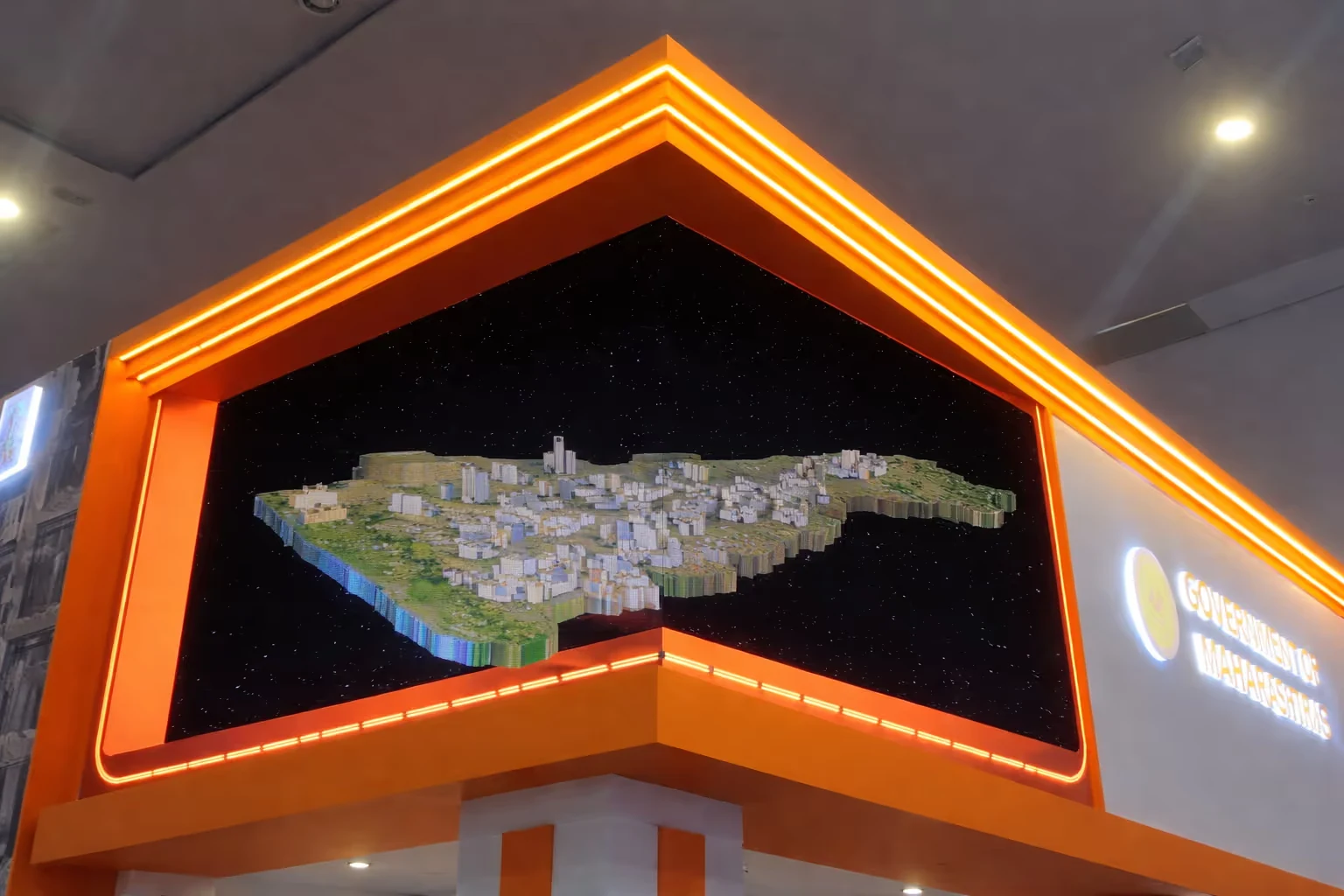 3D model of a city landscape displayed on a corner screen with orange neon lighting, inside a building with 'Government of Maharashtra' signage.