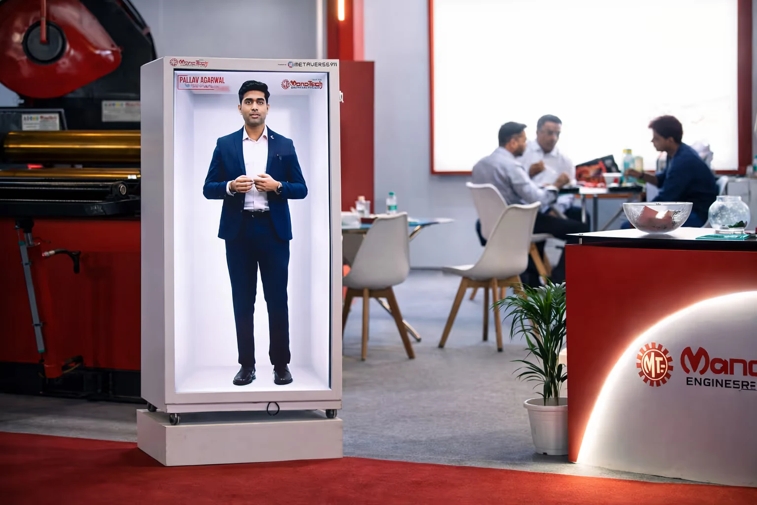 Digital life-sized display of a man in a suit inside a white rectangular frame, at an industrial-style exhibition booth with a few people sitting and talking in the background.