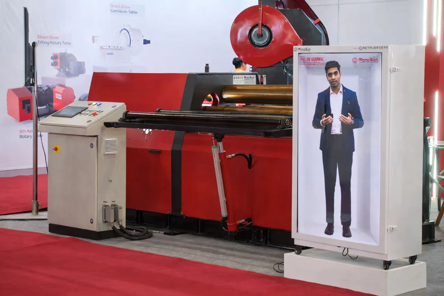 Holographic display of a man in a suit presenting, placed next to a red and black industrial machine with a control panel.