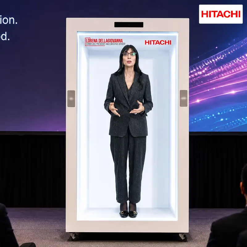 A Hitachi executive appearing as a full-height hologram on a HoloBox911 stage unit at the Hitachi corporate event in New Delhi, with the on-screen name plate and HITACHI logo visible