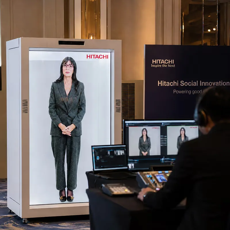 HoloBox911 holographic display unit on stage in front of a Hitachi Social Innovation Powering good backdrop, with a crew member alongside, at the Hitachi corporate event in New Delhi