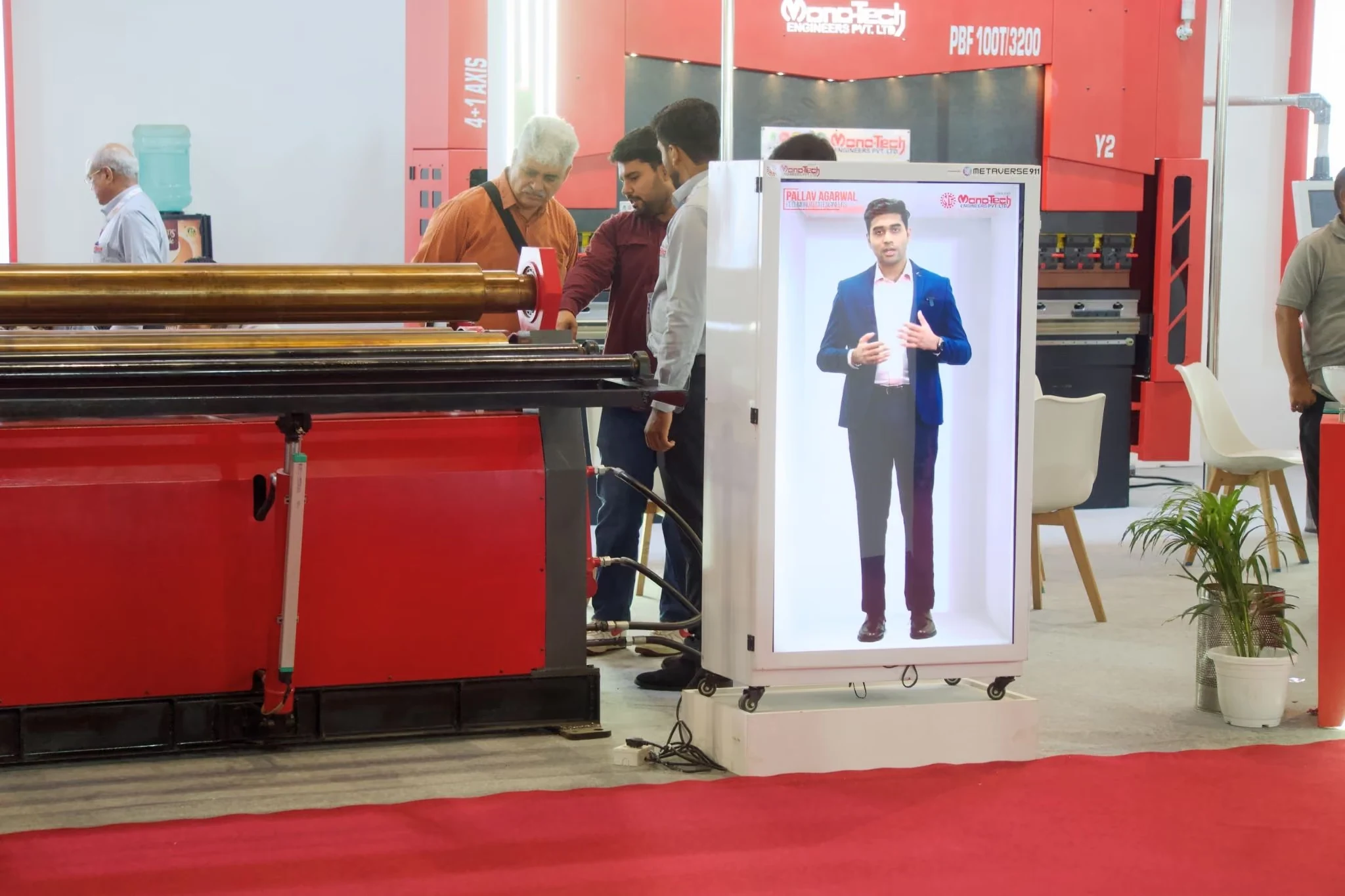 Visitors at the Monotech Engineers stand at IMTOS 2025, with the HoloBox911 unit playing the recorded welcome beside red sheet metal machinery on the show floor.