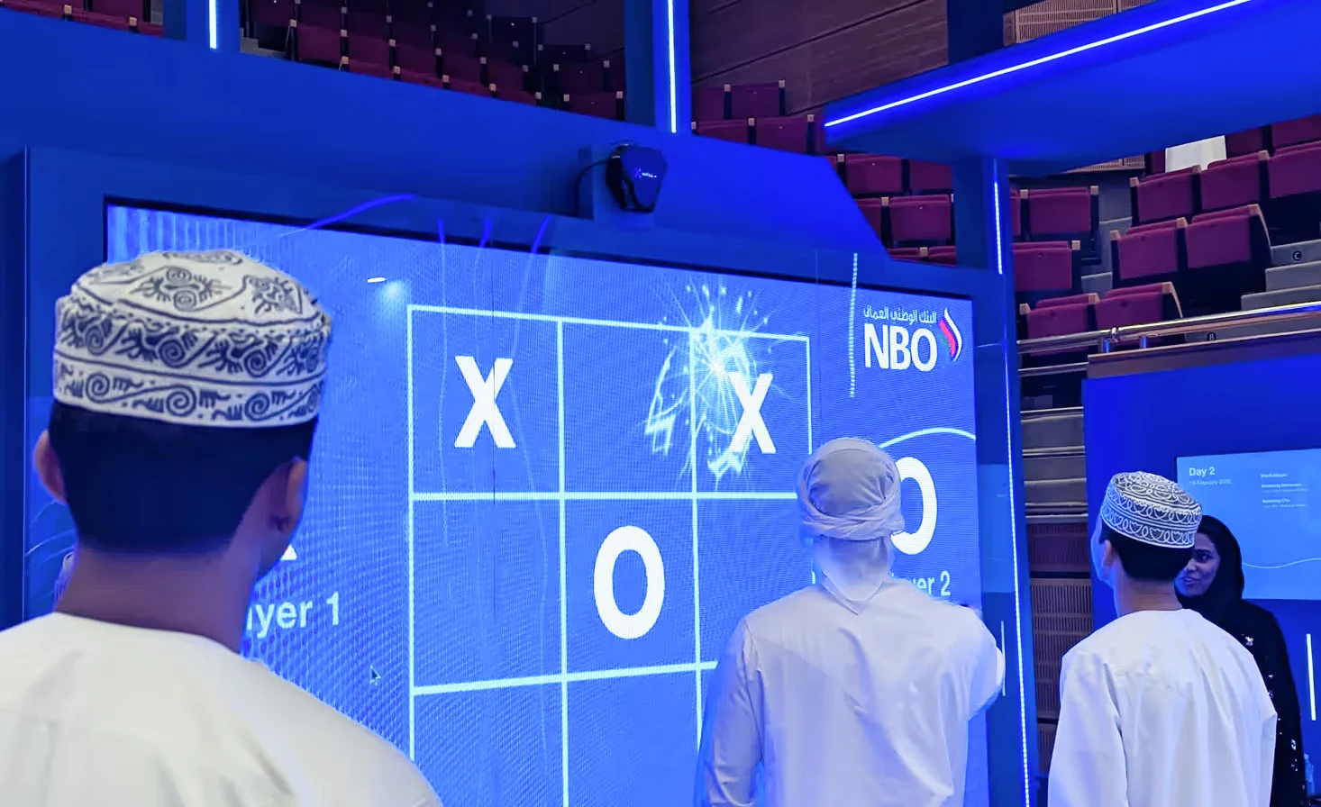 Three visitors in traditional Omani attire playing a multiplayer tic-tac-toe game on the NBO-branded XWall interactive screen at an exhibition.